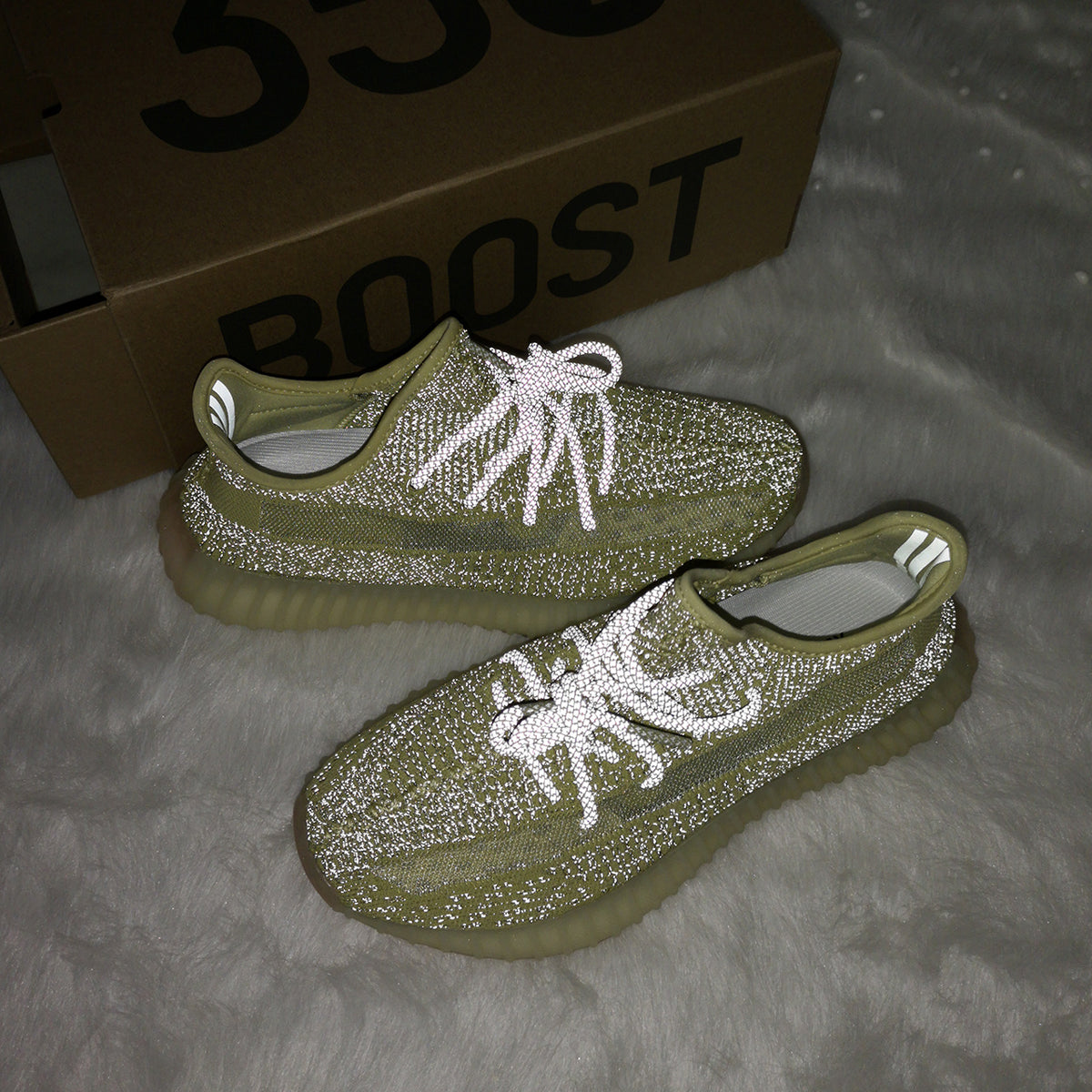 Lifestyle photo of olive green sneakers with reflective-looking laces beside a box visibly marked BOOST.