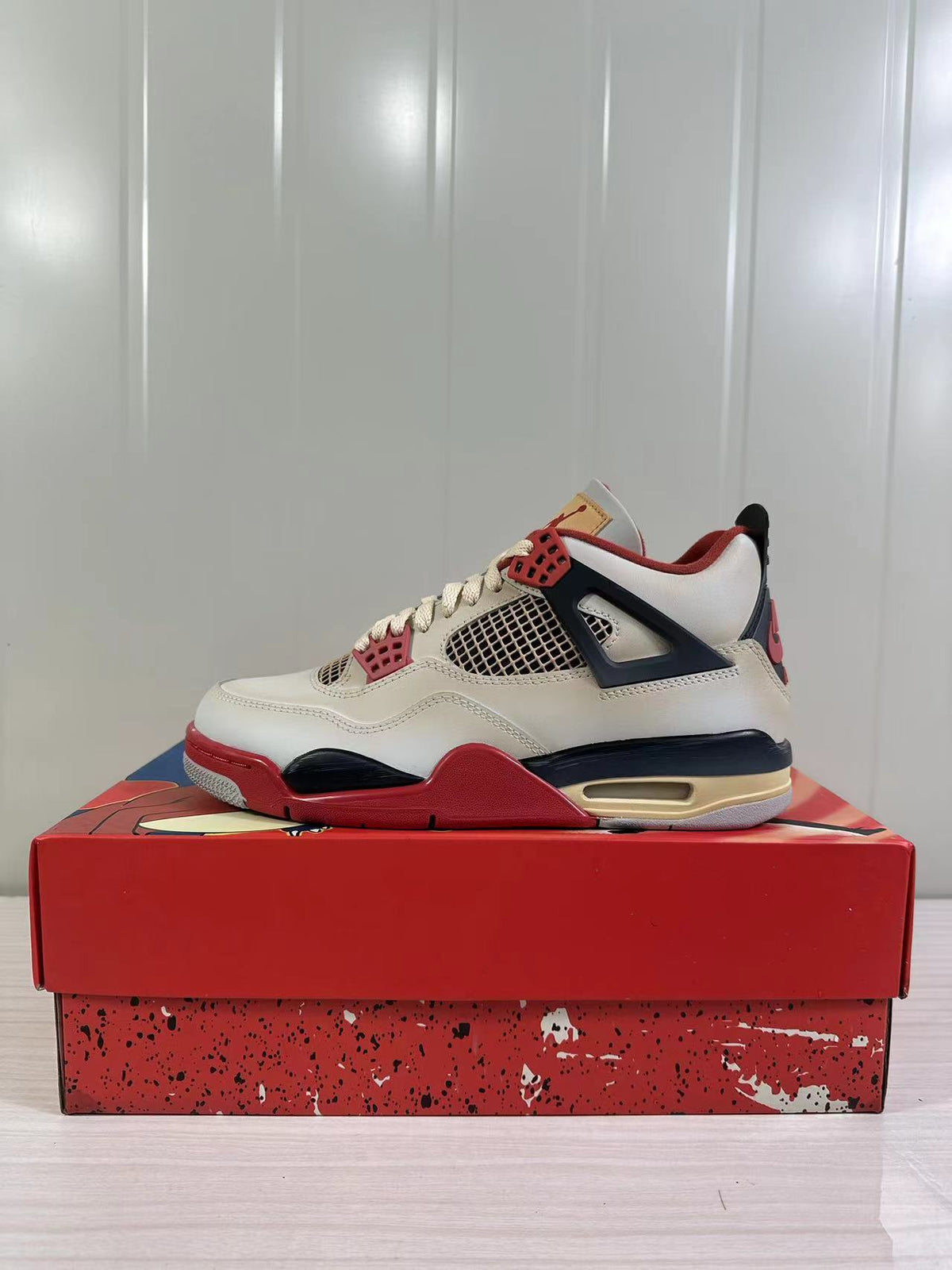 Side view of off-white Jordan Air Jordan 4 Retro sneakers with red and dark navy accents on a red shoebox.