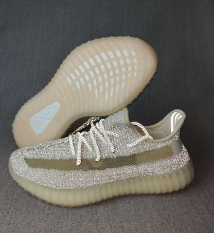 Side and outsole view of silver-toned Yeezy 350 V2 sneakers with an olive stripe and translucent cream sole.