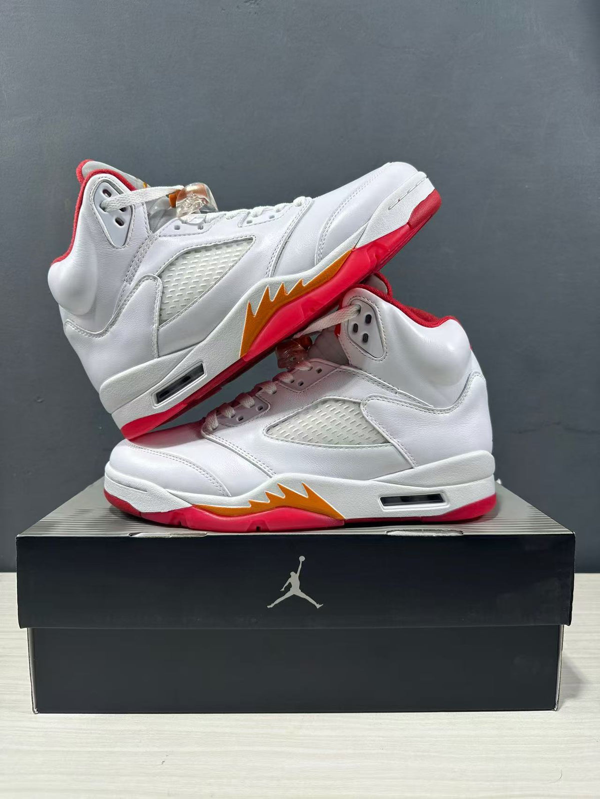 Pair of white high-top sneakers arranged on a Jordan shoe box, showing perforated side panels, red soles and orange accents.