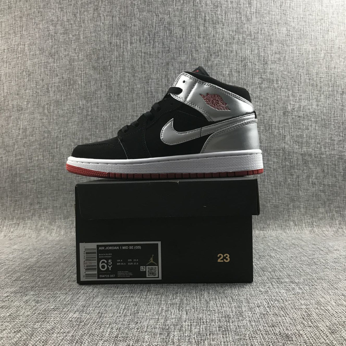 Side profile of one sneaker placed on its box; the box label shows Air Jordan 1 Mid SE GS, Black/Silver, size 6.5Y, EU 37.5 and style code 554725-057.