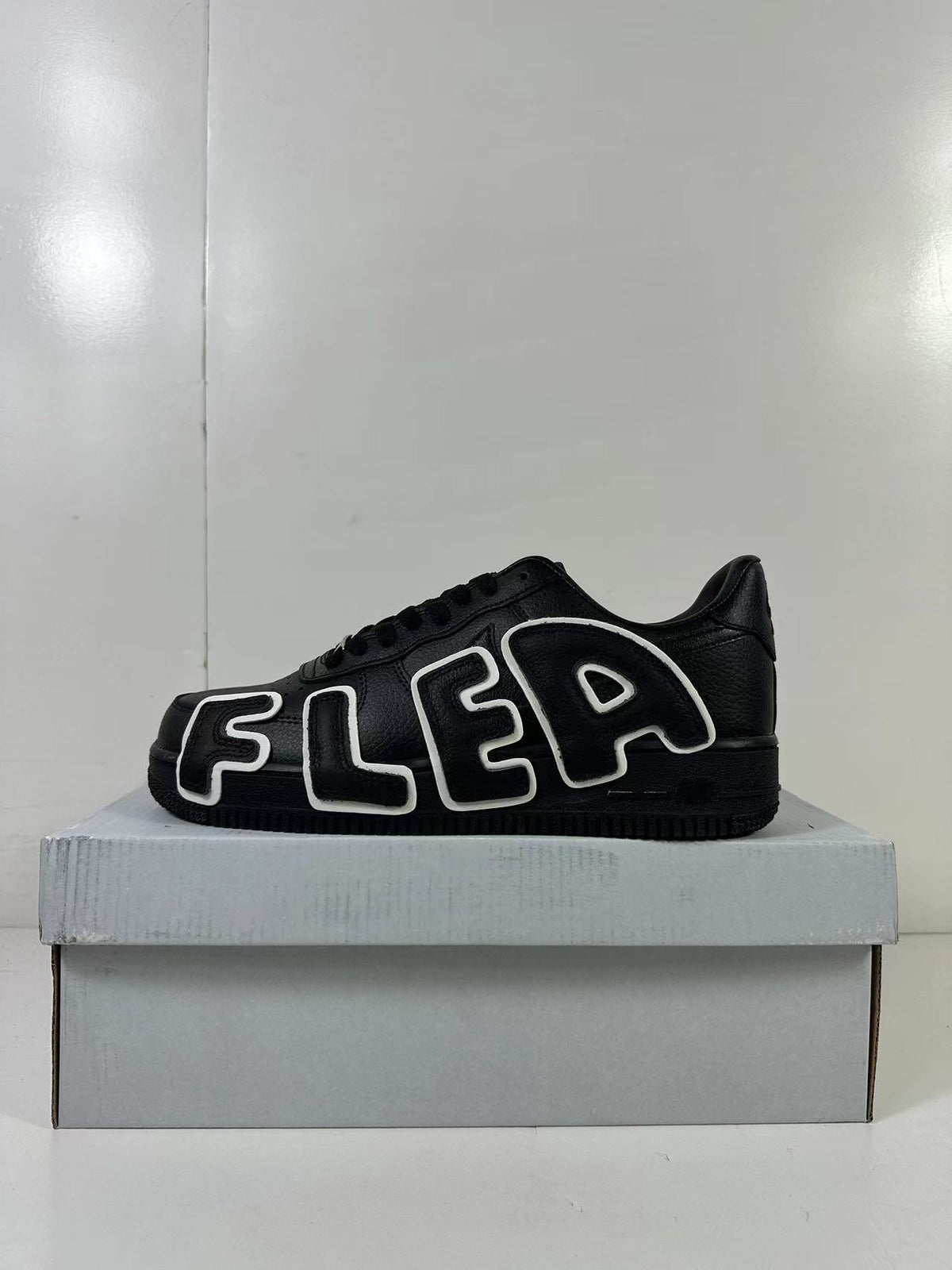 Side view of a black low-top sneaker with large outlined FLEA lettering in black and white.