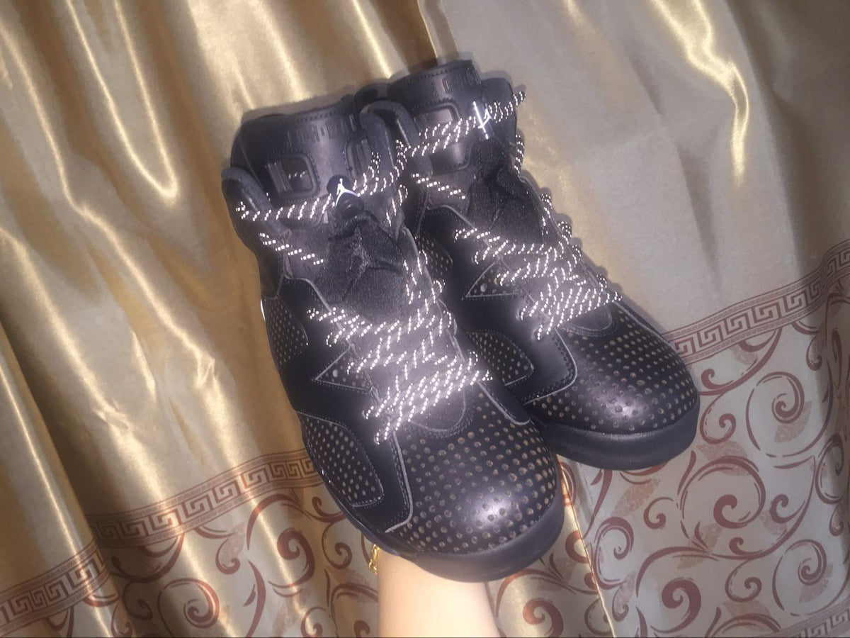 Pair of black sneakers displayed against a patterned curtain, with reflective-style laces catching the light.