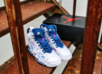 Pair of white high-top Jordan sneakers with blue laces, pink branding, and multicolor side panels displayed beside a shoebox.