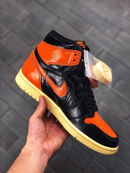 Handheld angled view showing the orange toe panel, black upper, orange collar, and spare laces in a clear bag.