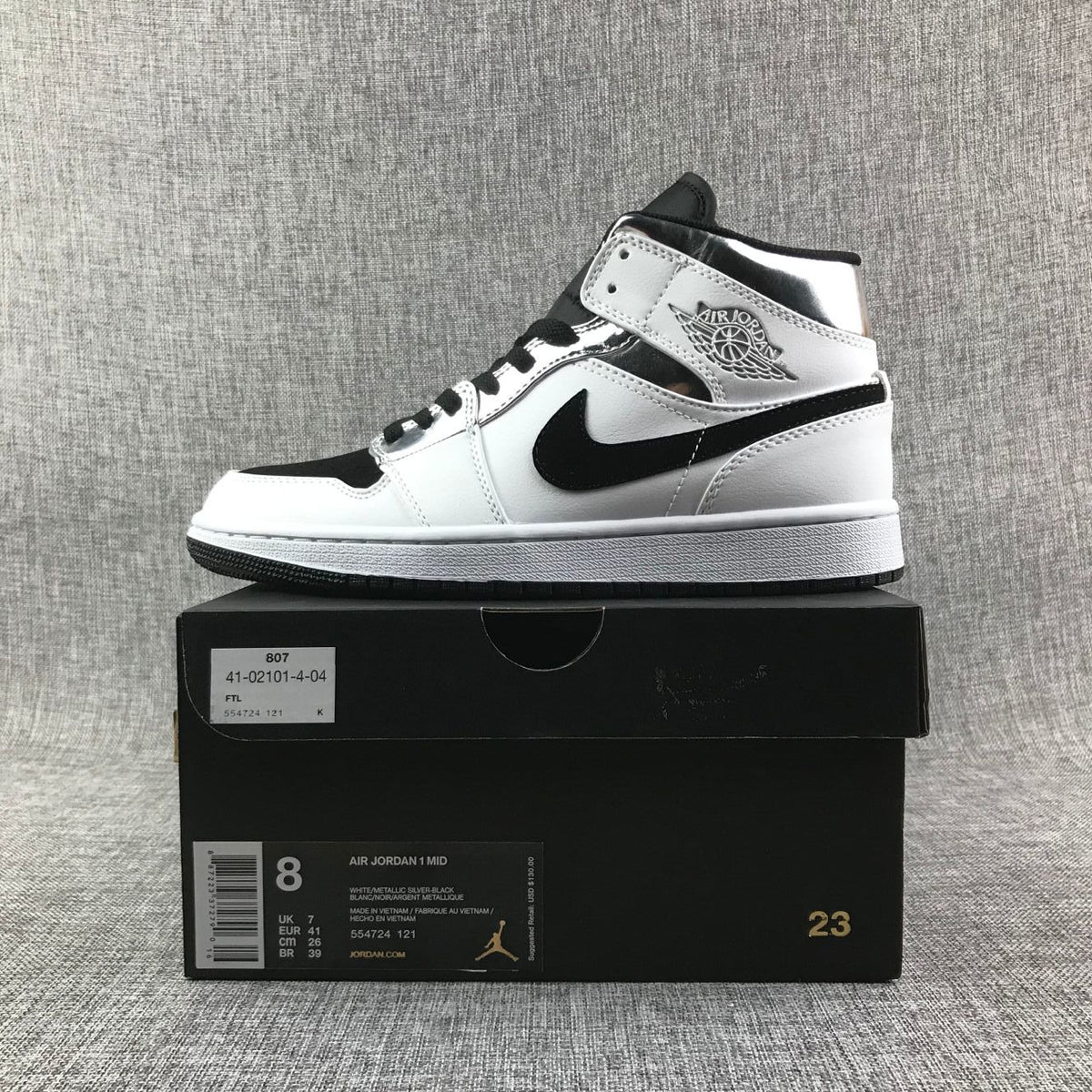 Side view of the sneaker positioned on its box; the visible label lists Air Jordan 1 Mid, EU 41, and the colors white, metallic silver, and black.