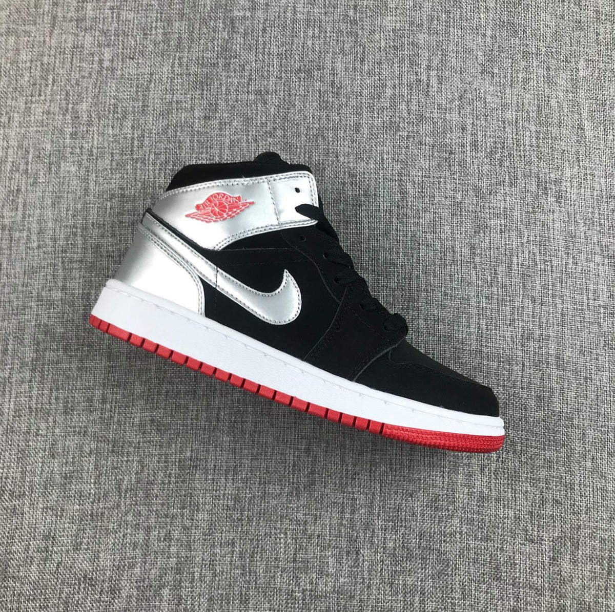 Side view of a black and silver Air Jordan 1 Mid SE GS sneaker with a white midsole and red outsole.