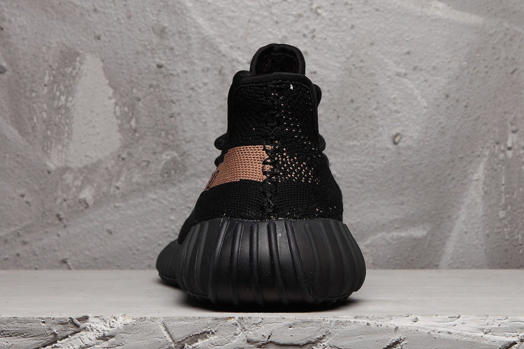 Rear view of a black Yeezy 350 V2-style sneaker with a ribbed black outsole and peach-toned stripe wrapping toward the heel.