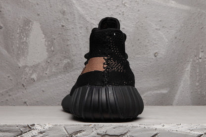 Rear view of a black Yeezy 350 V2-style sneaker with a ribbed black outsole and peach-toned stripe wrapping toward the heel.