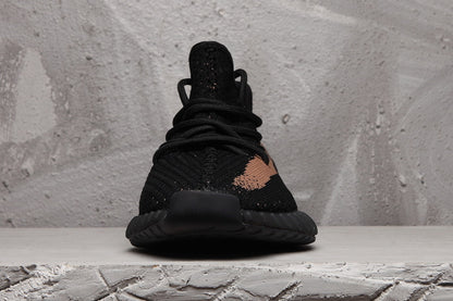 Front view of a black Yeezy 350 V2-style sneaker with black laces, textured upper, and a peach-toned side detail.
