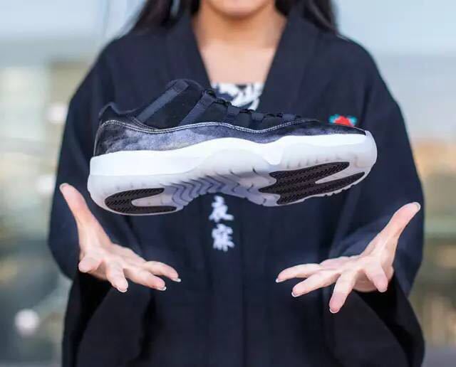 Person holding one sneaker to display its black and silver upper, white midsole, and outsole pattern.