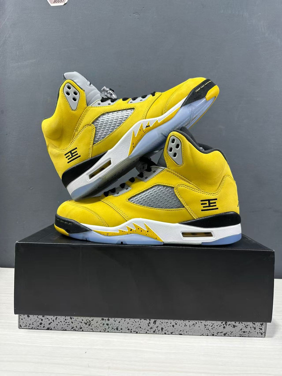 Two yellow sneakers arranged on a black box, showing their side panels and translucent outsoles.
