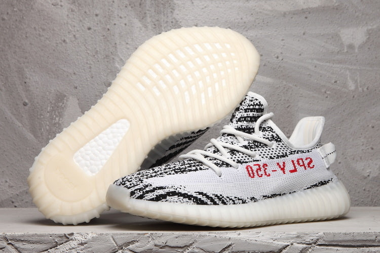Side view of black-and-white Yeezy 350 V2 sneakers with a light ribbed sole; one shoe is lifted to show the outsole.