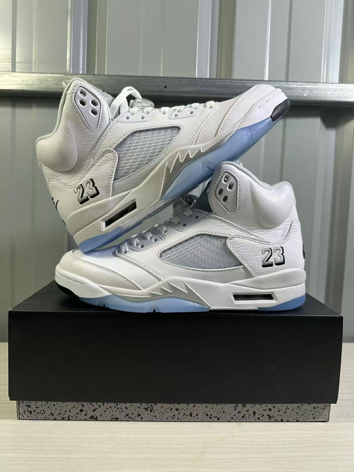 Two white and silver high-top sneakers arranged on a black shoe box from an angled side view.