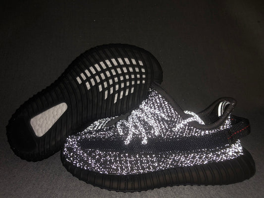 Black Yeezy 350 V2 style sneakers shown with reflective detailing illuminated by flash.