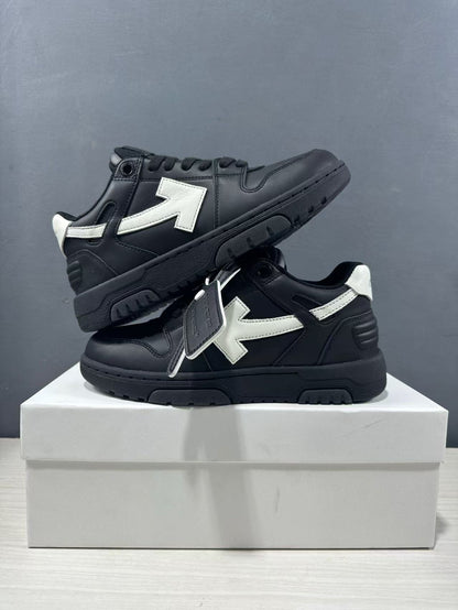 Two black and white sneakers stacked on a box, showing the contrasting arrow panels, heel construction, and attached product tag.